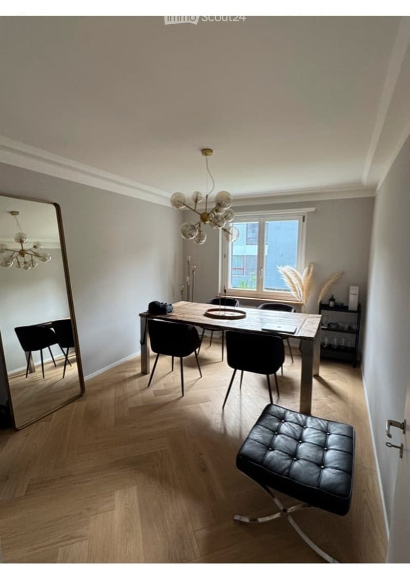 Modern dining area with a wooden dining table, black chairs, a black leather stool, and a large mirror on the wall. Natural light streams in from a window, and a decorative chandelier hangs from the ceiling.