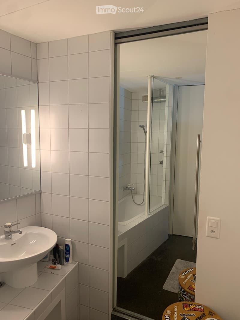 Modern bathroom with white tiled walls, a sink with a modern faucet, a bathtub with a glass shower enclosure, and toiletries on the sink counter.