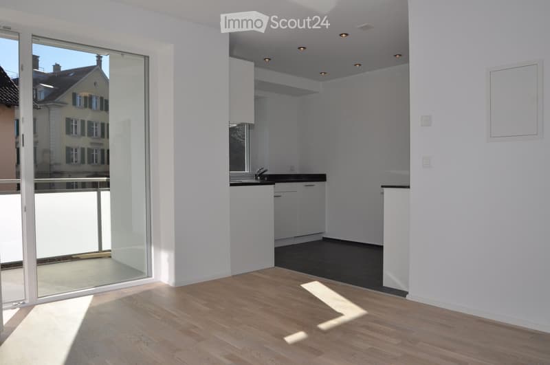 Empty room with wooden flooring, white walls, and a small kitchen area with cabinets and a sink. Large glass sliding doors lead to a balcony with a view of a neighboring building.