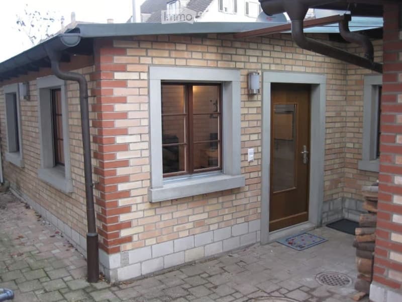 Exterior view of a small brick building with a flat roof, featuring a wooden door, a window with a view into a room, and a paved stone pathway.