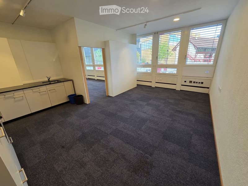 A small, furnished room with a sink, cabinets, and trash bins. The room has carpeted flooring and large windows with blinds.