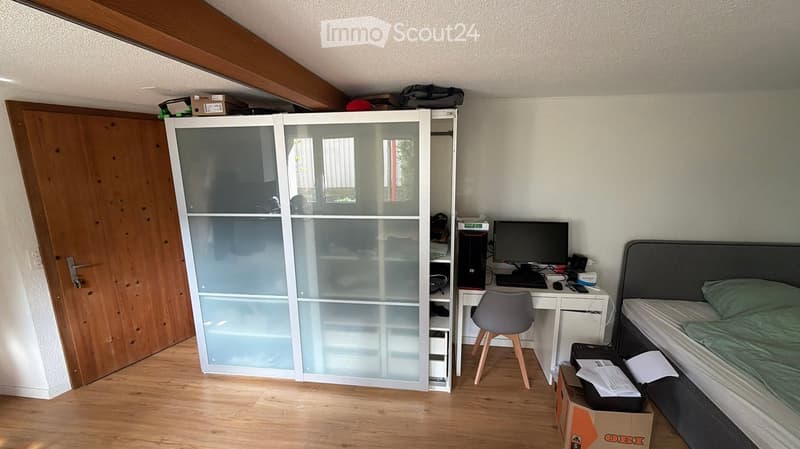 A furnished bedroom with a bed, desk, chair, computer, wardrobe, and a wooden door.