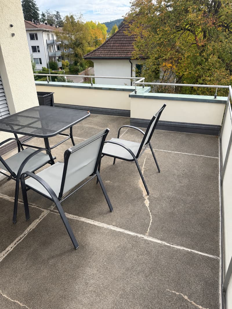 Balcony with a black metal table and two matching chairs, overlooking a residential area with trees and neighboring buildings.