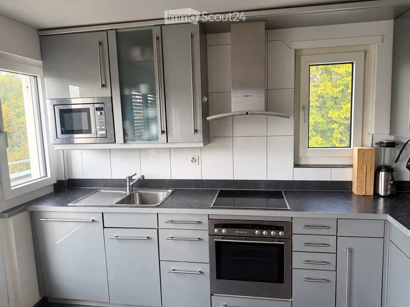 Modern kitchen with grey cabinets, stainless steel appliances, and a black countertop. Features include a microwave, oven, sink, faucet, blender, and cutting board.
