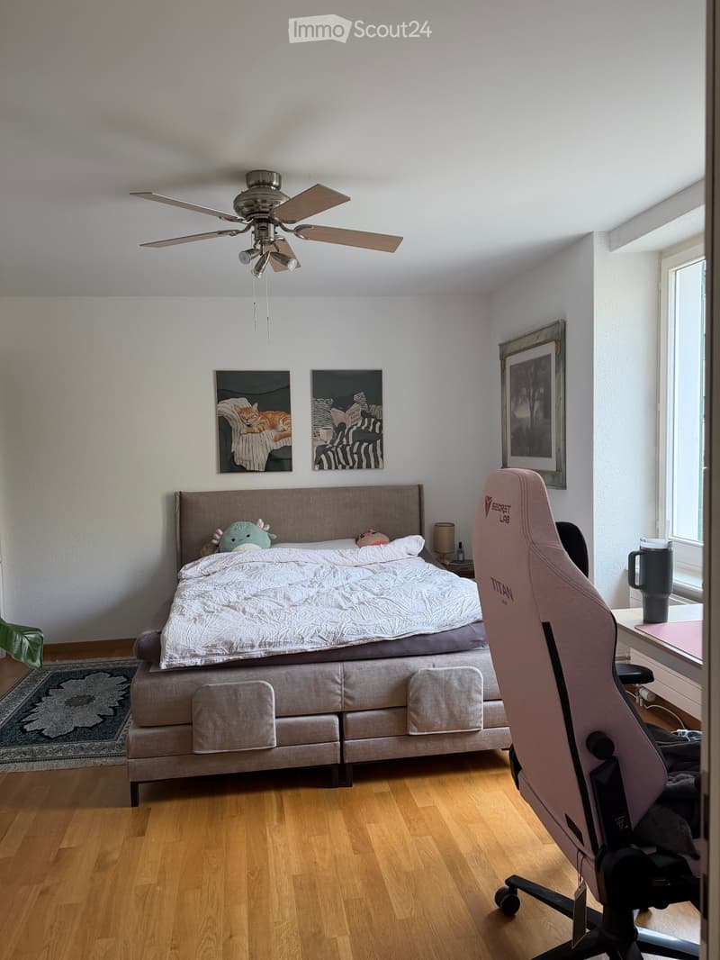 A well-lit bedroom with a large bed, a gaming chair, a small table with a lamp, and several picture frames on the wall.