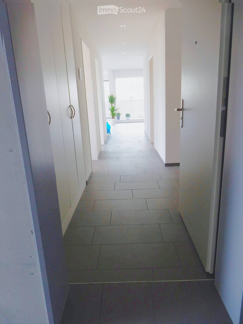 Clean, modern hallway with white walls, tiled flooring, and several doors on both sides. Natural light streams in from the end of the corridor.