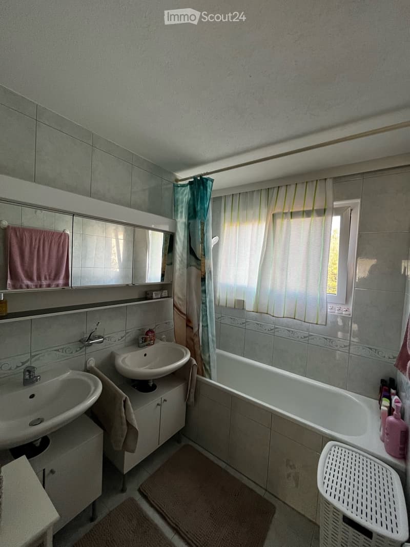 Bathroom with two sinks, a bathtub, a mirror, and a window with a shower curtain. Includes a towel, a bathroom cabinet, and a laundry basket.