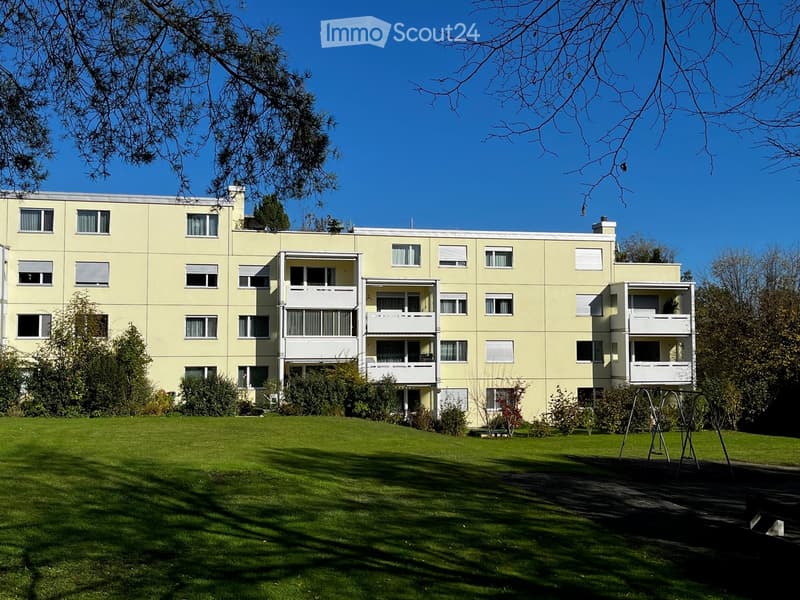 A multi-story residential building with light yellow walls, multiple windows, and balconies, surrounded by a well-maintained lawn and trees.