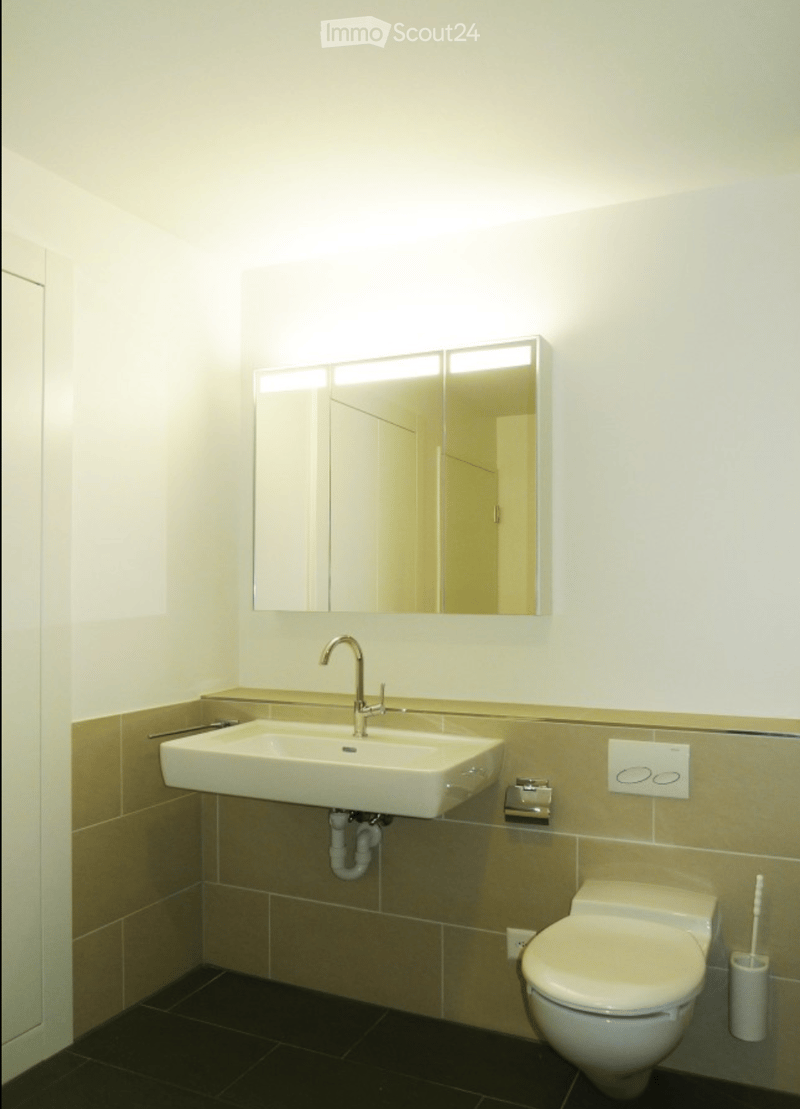 Modern bathroom with a white sink, chrome faucet, large mirror, toilet, and toothbrush holder. Beige tiled walls and dark tiled floor.