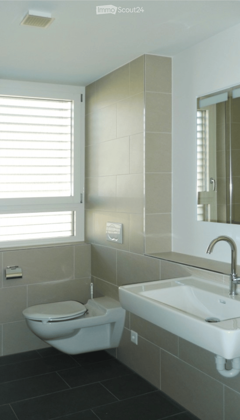 Modern bathroom with a toilet, sink, and mirror. Features tiled walls and a window with blinds.