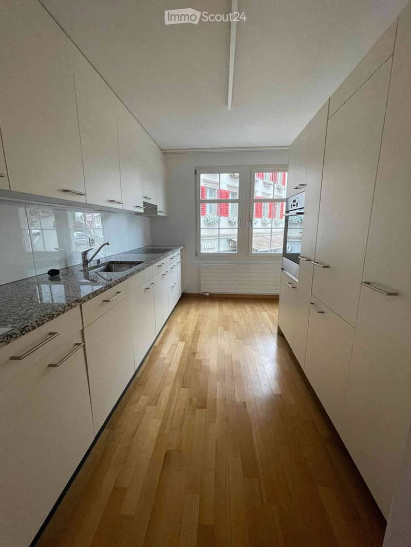 Empty kitchen with light-colored cabinets, granite countertop, wooden floor, and large windows showing a view of a building with red shutters.