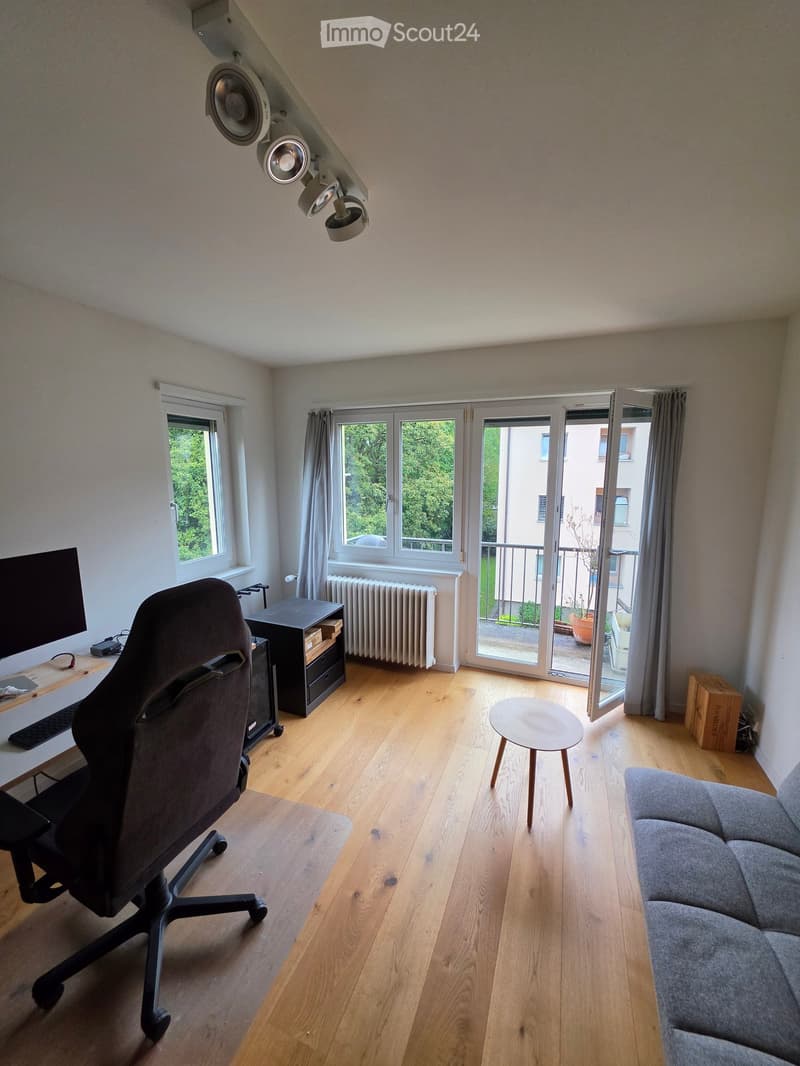 A modern, well-lit room with wooden flooring, large windows, and a balcony door. Features include a grey sofa, a round wooden stool, a desk with a screen and chair, and a radiator under the windows.