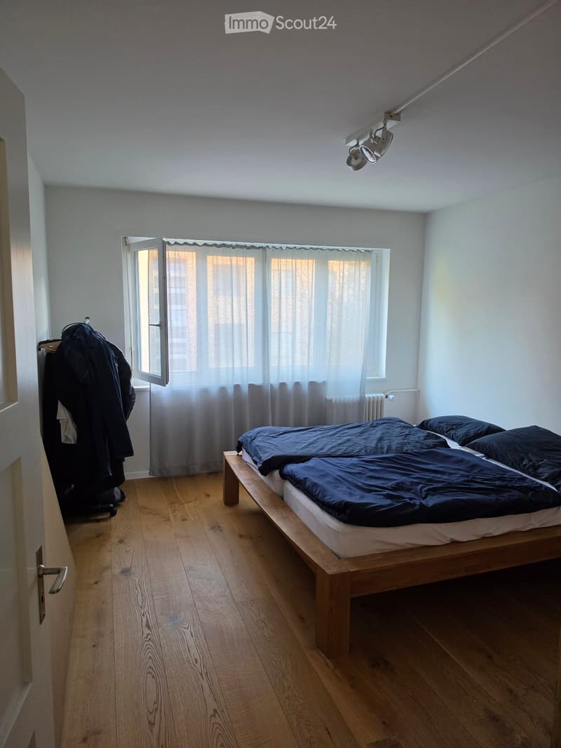 A modern bedroom with wooden flooring, a single bed with dark blue bedding, a window with white curtains, a radiator, and a wardrobe with hanging clothes.
