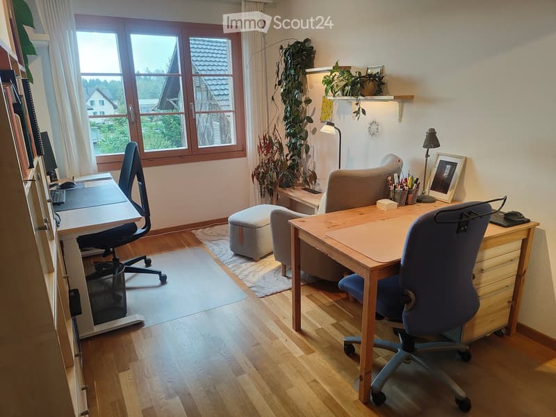 Well-lit study or home office with wooden flooring, large windows, two desks, ergonomic chairs, a small sofa, a stool, a floor lamp, and decorative plants.