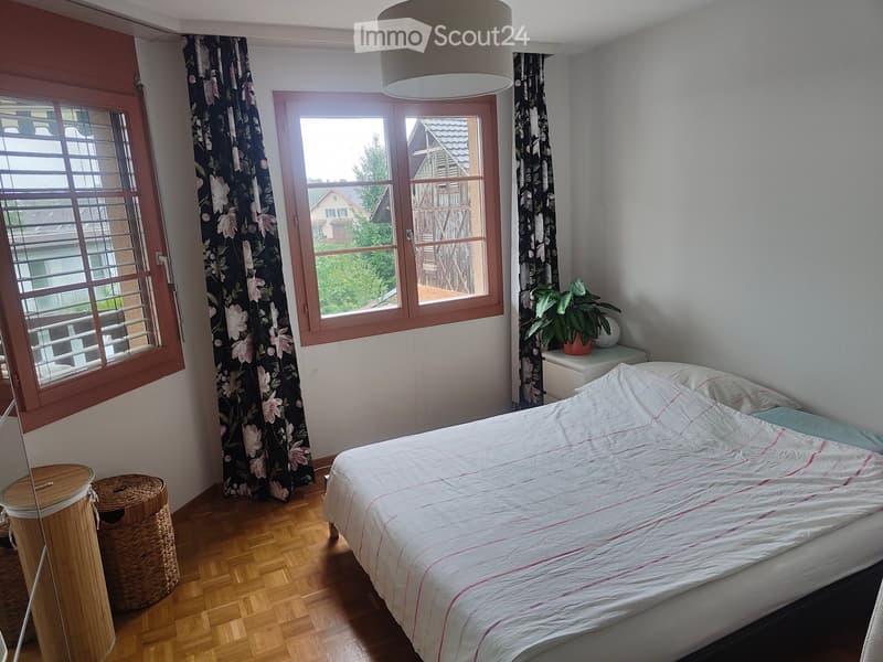 Cozy bedroom with wooden flooring, large windows with floral curtains, a neatly made bed with white and pink bedding, a nightstand with a potted plant, and a wicker stool near the window.