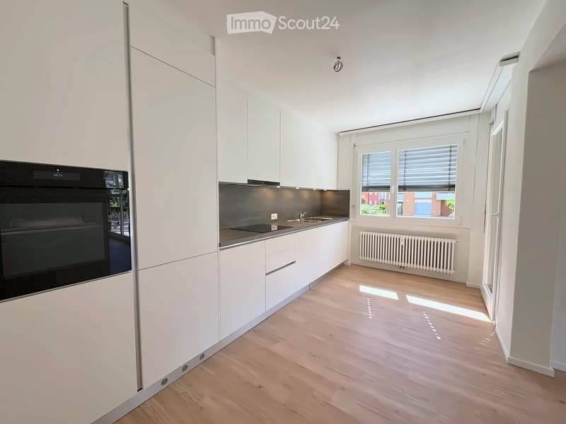 Modern kitchen with white cabinets, wooden flooring, and a black oven. Features a sink, radiator, and large windows with blinds.