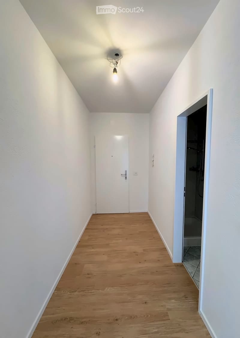 Empty hallway with wooden flooring, white walls, and a single light fixture on the ceiling. A closed door is visible at the end of the hallway, and an open doorway on the right leads to a tiled bathroom area.