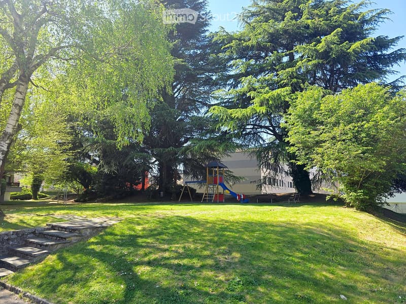 A well-maintained garden with a grassy lawn, stone steps, and a playground featuring a slide and climbing frame, surrounded by tall trees and a building in the background.
