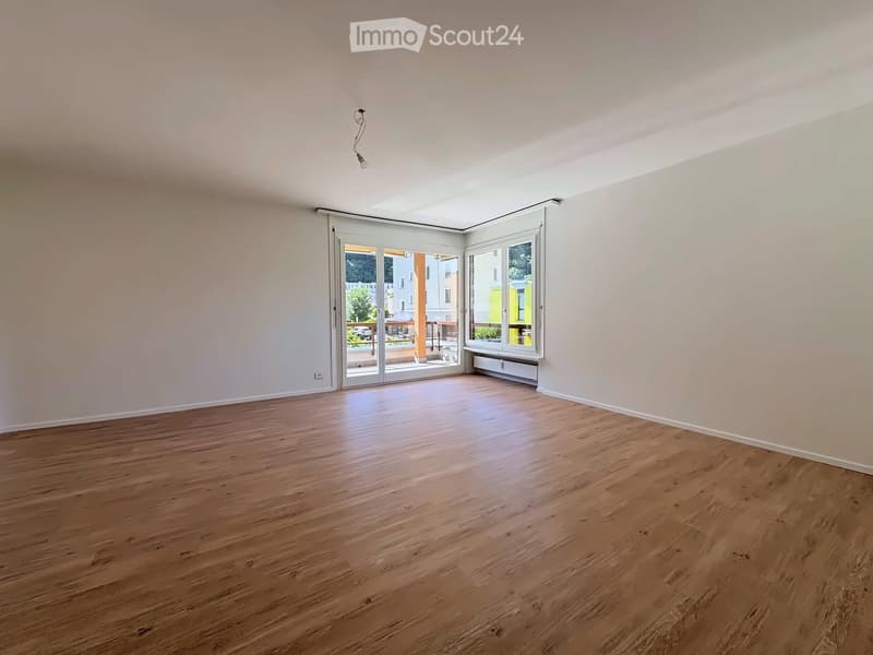 Empty room with wooden flooring, white walls, large windows, and a sliding glass door leading to a balcony.