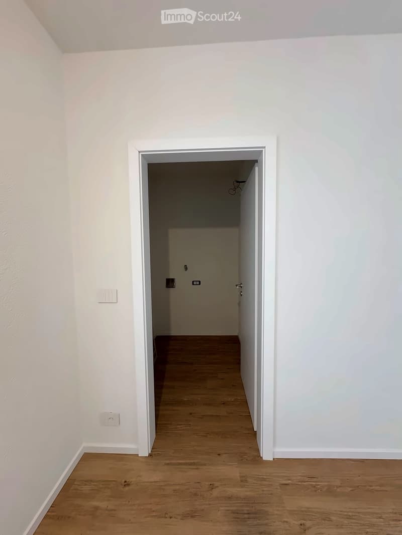 Empty hallway with light wooden flooring and white walls, featuring a doorway leading to another room.