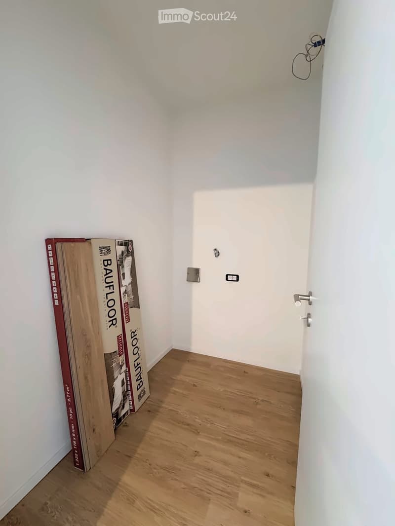 Empty hallway with wooden flooring, white walls, and a closed door at the end. There are some rolled-up flooring materials leaning against the left wall.