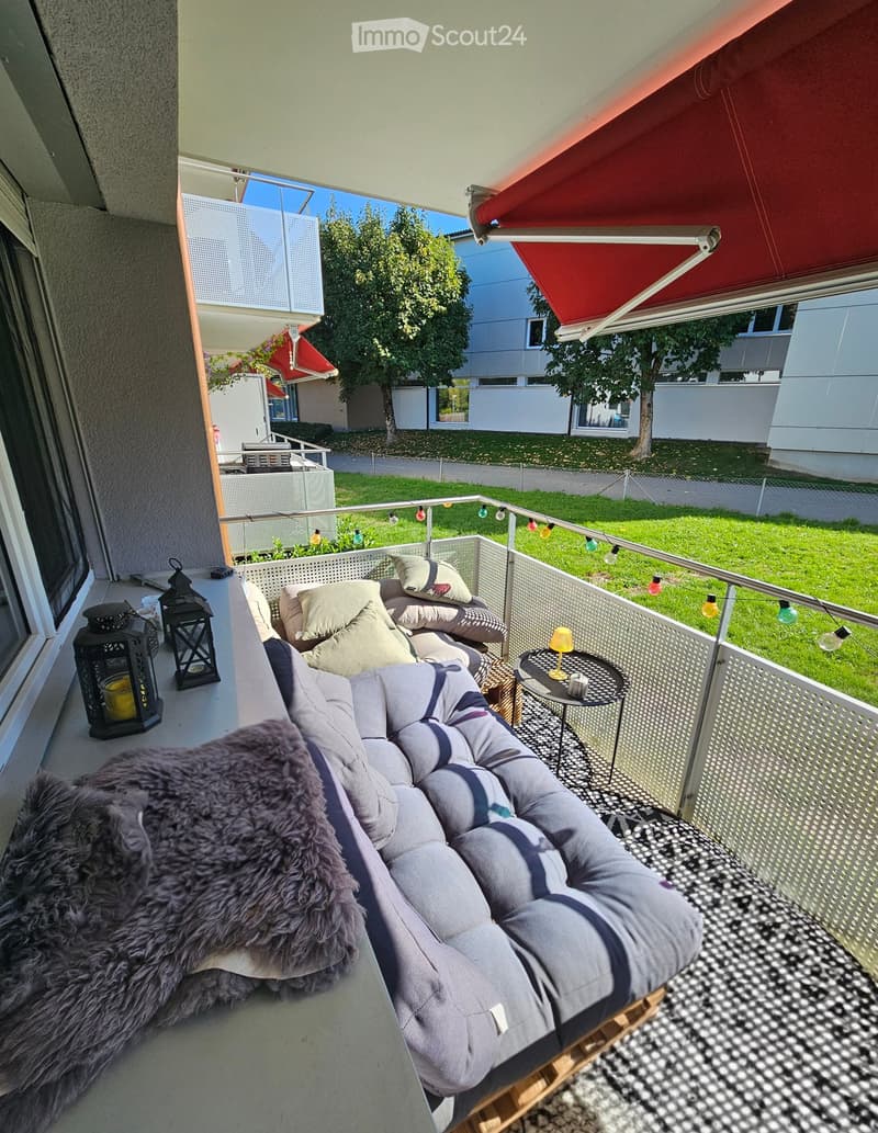Balcony with a grey sofa, small round table, and decorative lanterns, overlooking a green lawn and neighboring buildings.