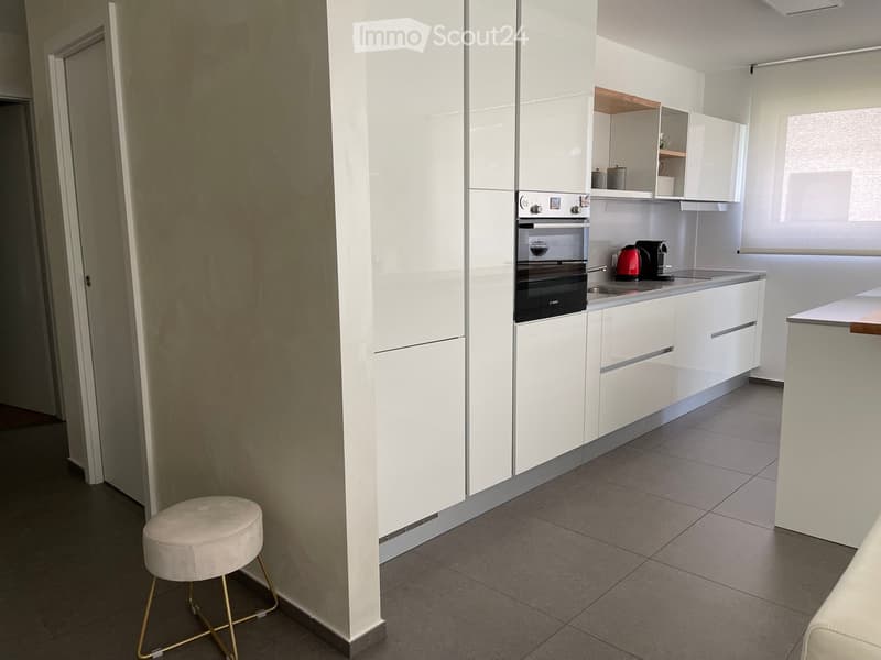 Modern kitchen with white cabinets, stainless steel oven, coffee maker, and a small stool near the entrance.