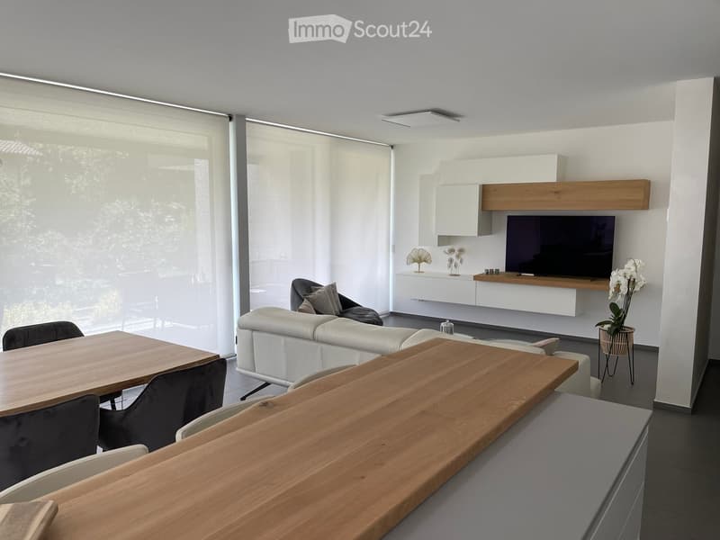 Modern living room with white walls, large windows with blinds, a white leather sofa, wooden coffee table, dining table with chairs, and a TV mounted on a white and wood entertainment unit.