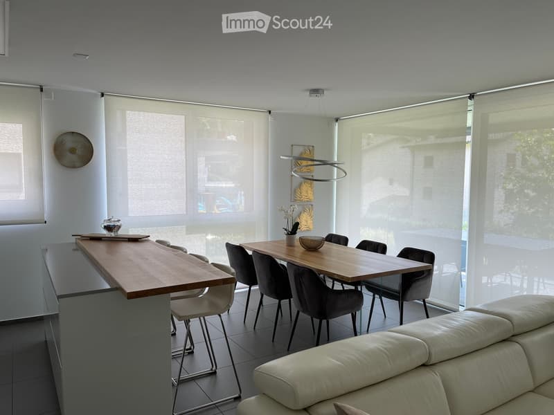 Modern living and dining area with large windows covered by roller blinds, a white leather sofa, a wooden dining table with six chairs, and a kitchen island with bar stools.
