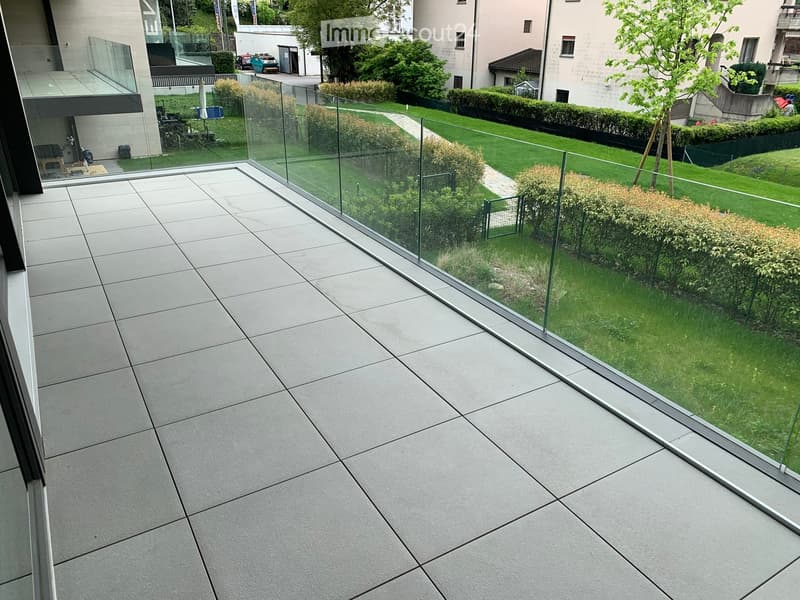 Modern balcony with large glass railing, tiled floor, and view of a well-maintained garden with green grass, bushes, and trees.