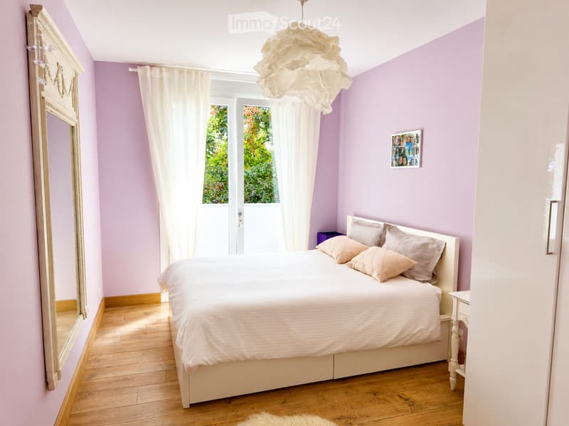 Cozy bedroom with light purple walls, wooden floor, white bed with pillows, nightstand, mirror, and a window with white curtains.