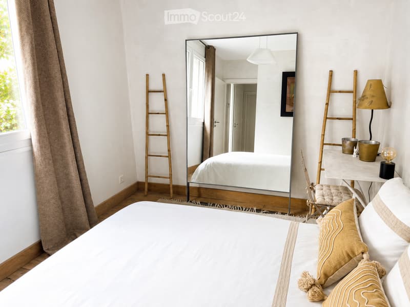A modern bedroom with a large bed, pillows, a mirror reflecting the room, a small table with a lamp, and a ladder leaning against the wall.