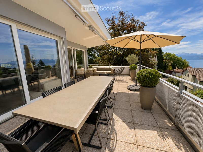 Balcony with a large table, chairs, umbrella, sofa, and potted plants, overlooking a scenic view of mountains and water.