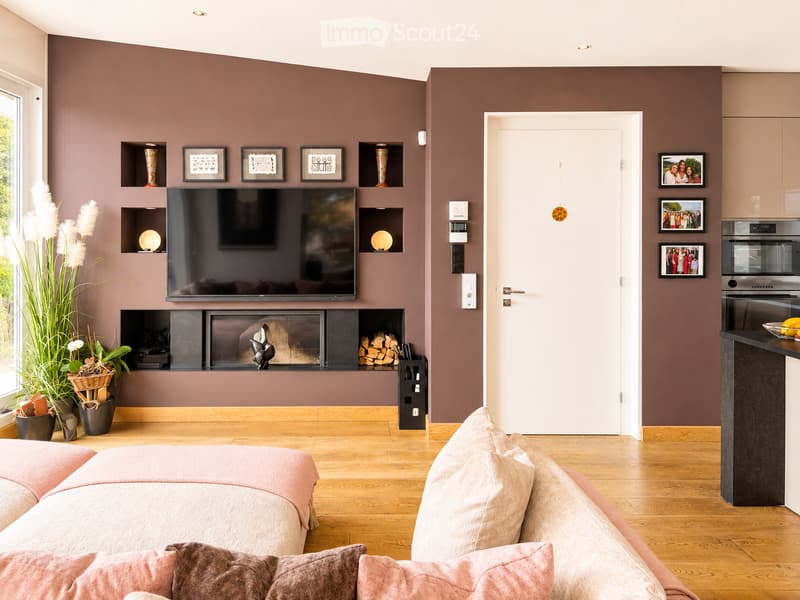 Modern living room with a fireplace, a large sofa, a television mounted on the wall, and built-in shelving with decorative items and framed pictures. The room features wooden flooring and a door leading to another area.