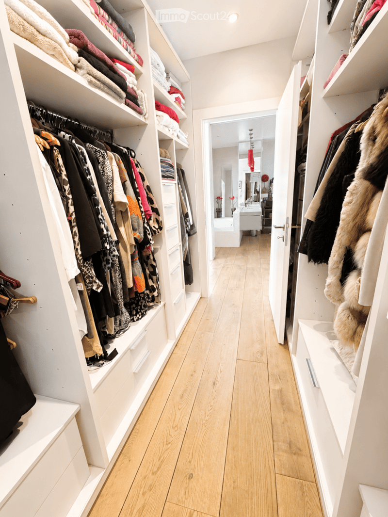 Spacious, well-lit hallway with built-in white shelving units filled with neatly organized clothing and accessories, leading to a bathroom area.