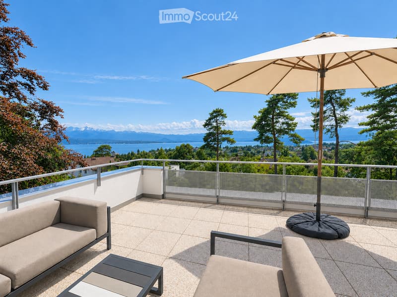 Balcony with a sofa, chair, table, and umbrella, overlooking a scenic lake and mountain view.