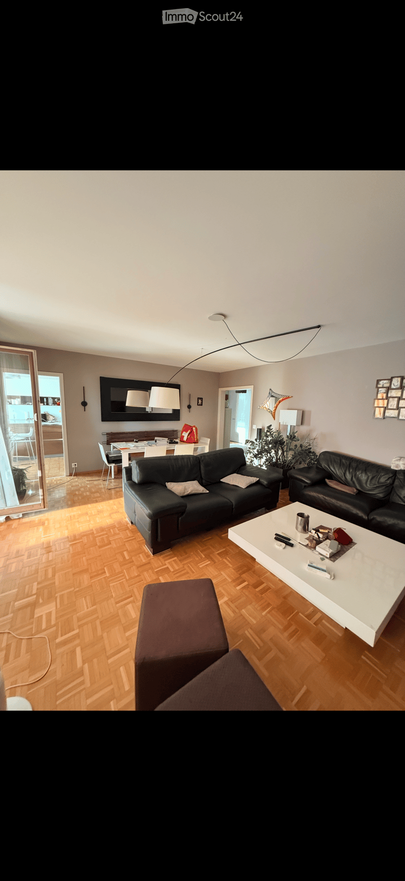 Modern living room with wooden flooring, black leather sofa, white coffee table, ottoman, and a dining area visible through an open doorway.