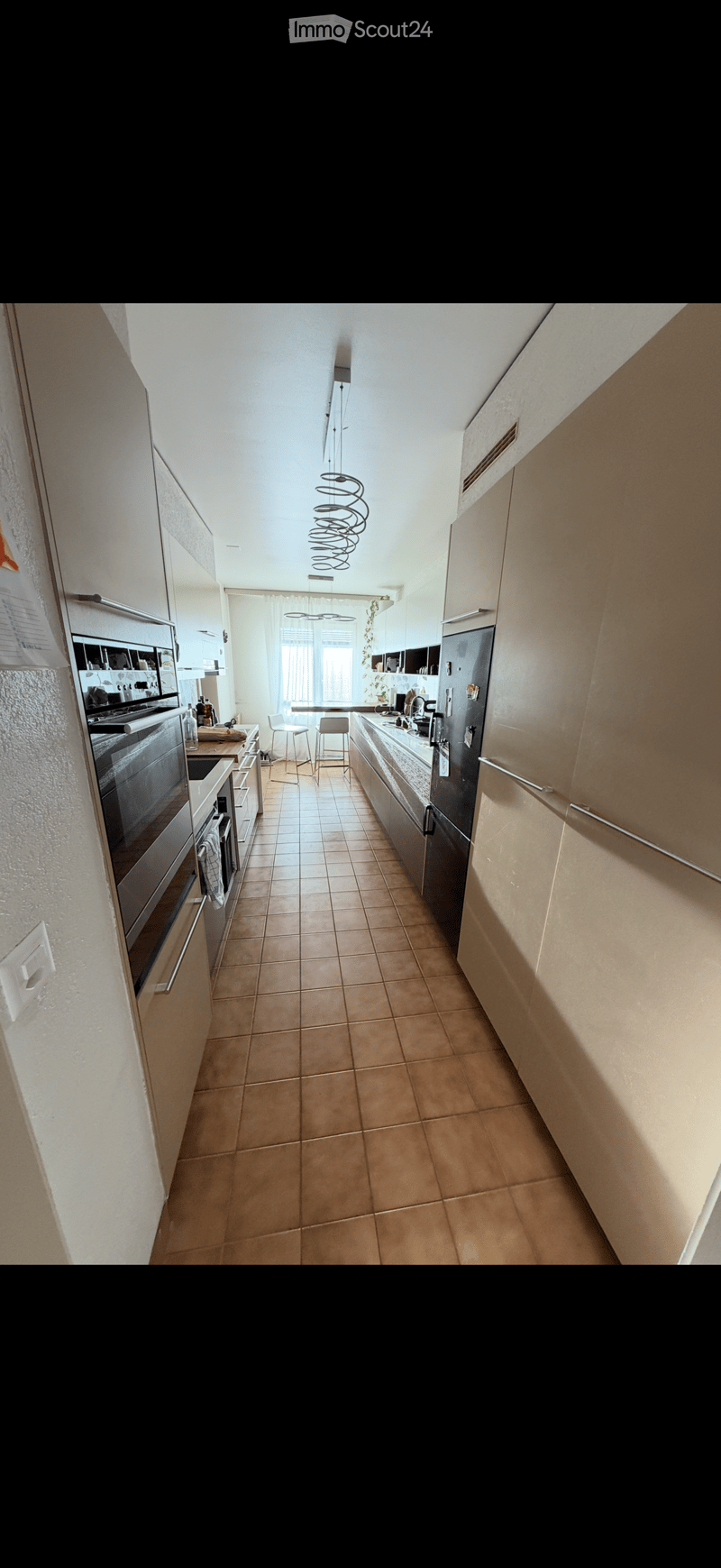 Modern kitchen with light-colored cabinets, stainless steel appliances, tiled floor, and two stools near the island.