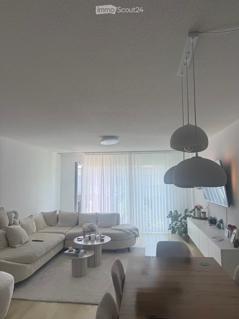 Modern living room with a large sectional sofa, coffee table, dining table, pendant lights, and a TV screen. The space features large windows with sheer curtains and a neutral color palette.