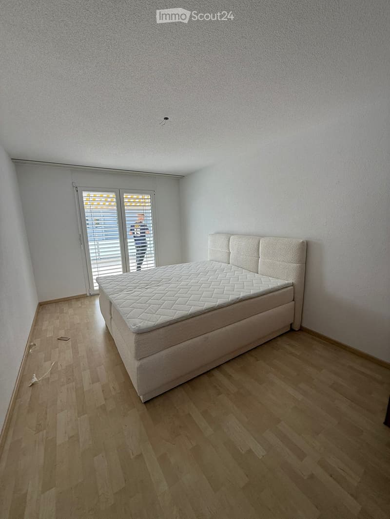 Minimalist bedroom with a single bed, headboard, and large window with blinds. Light wooden flooring and white walls.