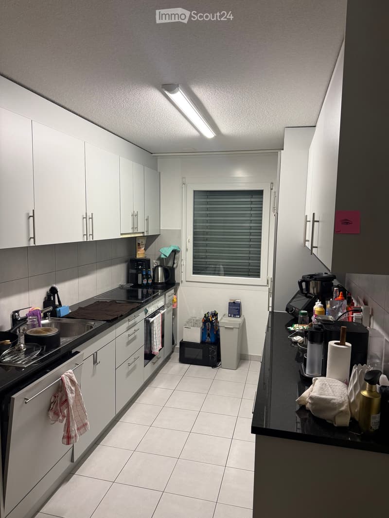 Modern kitchen with white cabinets, black countertops, and various appliances including a coffee maker, microwave, and toaster. The kitchen features a sink with a faucet, a stove, and a window with blinds.