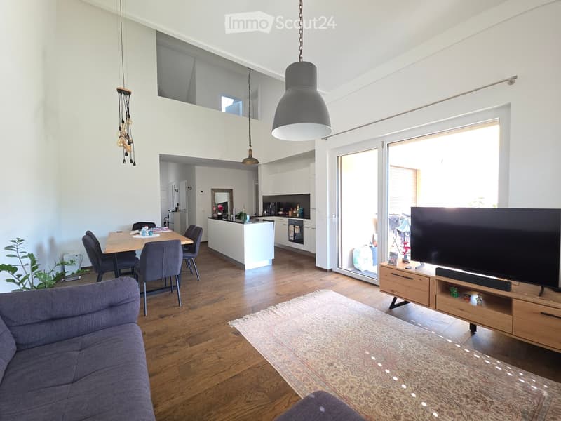 Open-plan living and dining area with a grey sofa, coffee table, dining table with chairs, kitchen island, and a television on a wooden stand. The space features wooden flooring, pendant lights, and large windows leading to a balcony.