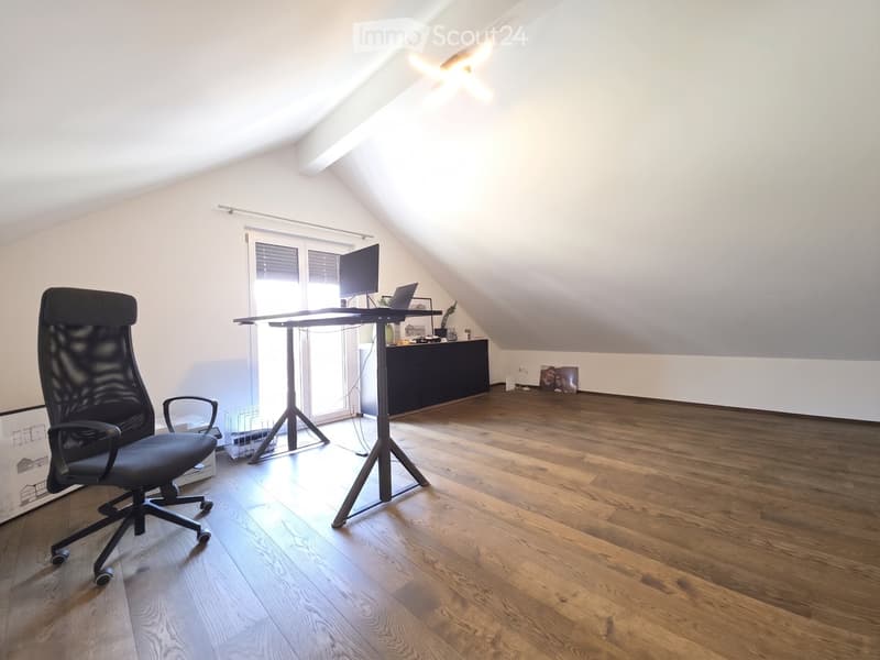 Bright attic room with wooden flooring, a standing desk, ergonomic chair, laptop, and a large window with blinds.