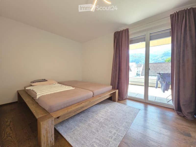 A modern bedroom with a wooden bed, pillows, blankets, a grey rug, and large windows with curtains leading to a balcony.
