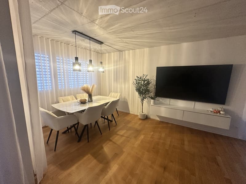 Modern dining room with a white dining table, six chairs, a large flat-screen TV mounted on the wall, pendant lights, and a potted plant. The room features wooden flooring and large windows with white curtains.