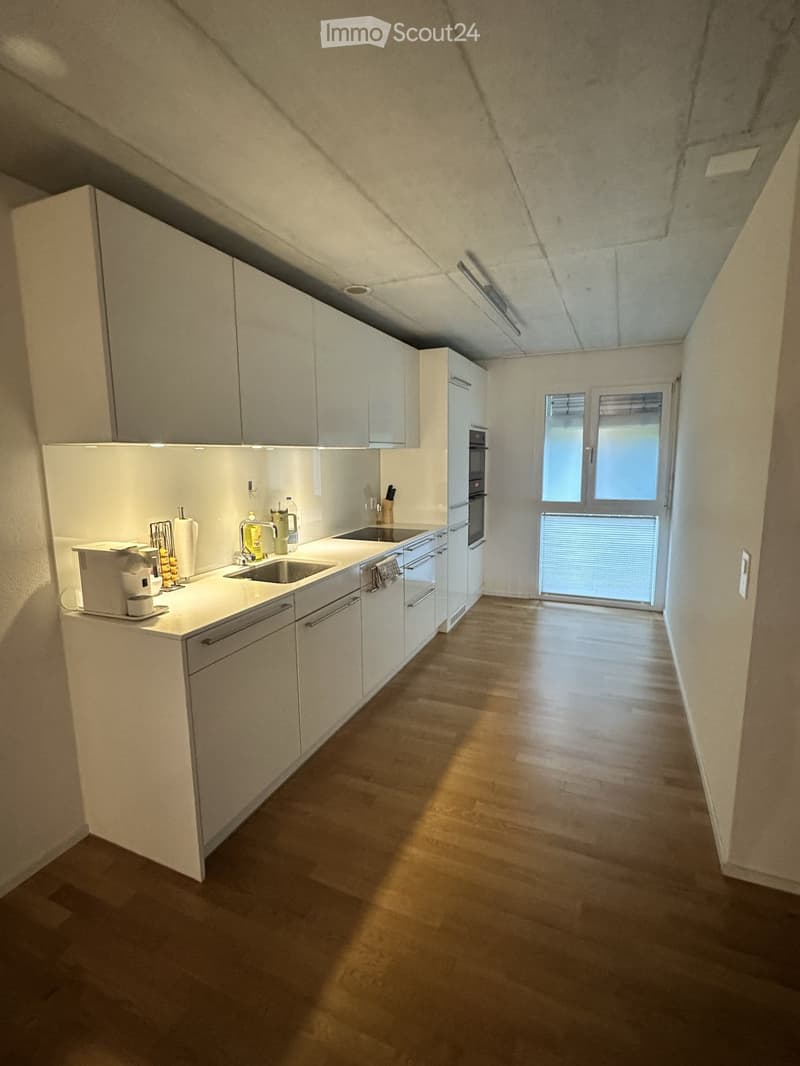Modern kitchen with white cabinets, stainless steel appliances, and a wooden floor. Features a sink, fridge, oven, and coffee machine on the counter.