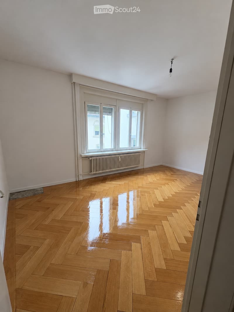 Empty room with polished wooden herringbone flooring, white walls, large window with blinds, radiator, and a single light fixture on the ceiling.