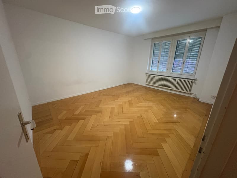 Empty room with wooden herringbone flooring, white walls, and large windows with shutters. A radiator is visible beneath the windows.
