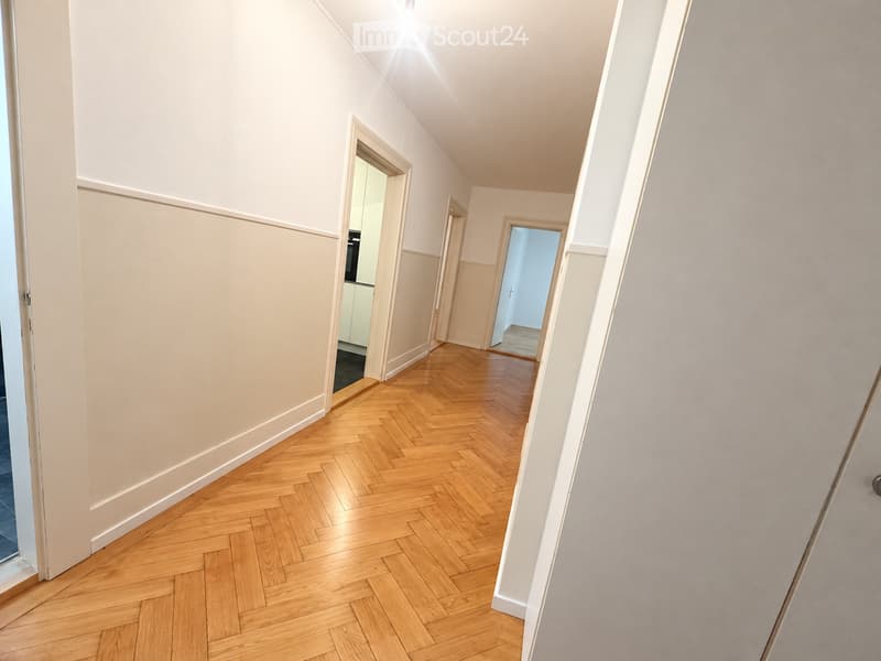 A narrow hallway with herringbone-patterned wooden flooring, light-colored walls, and several doors leading to adjacent rooms.