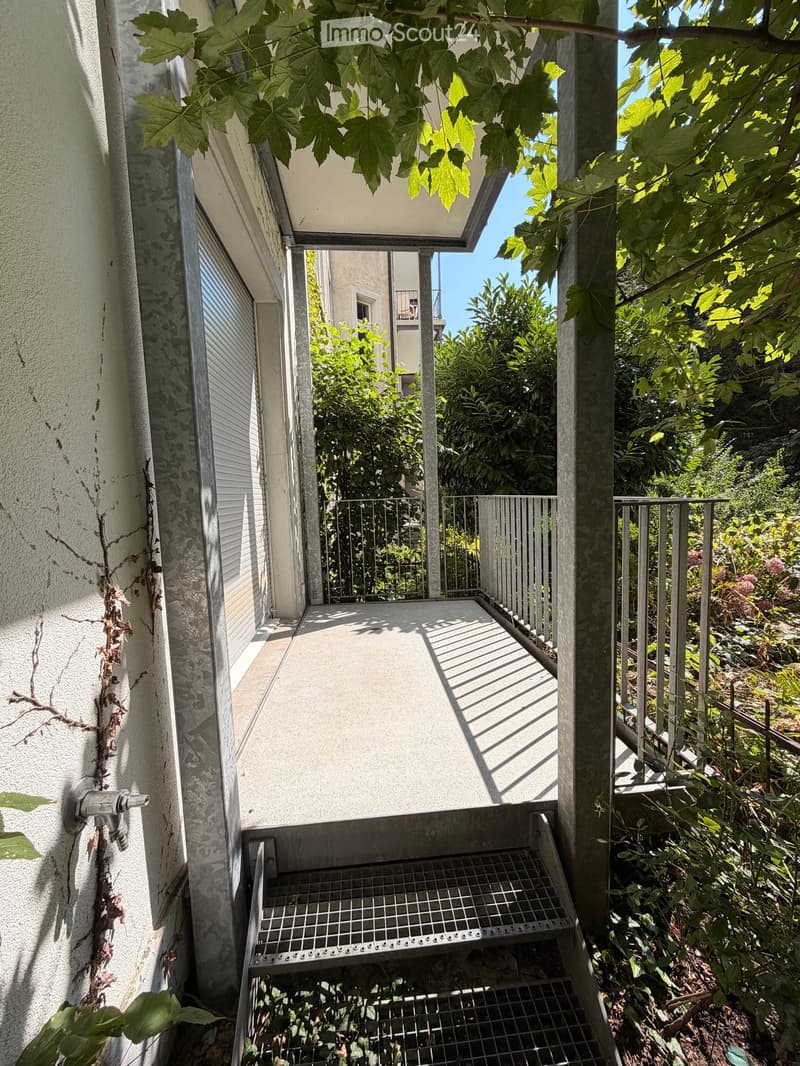 Balcony with metal railing, steps leading down, surrounded by greenery and trees.